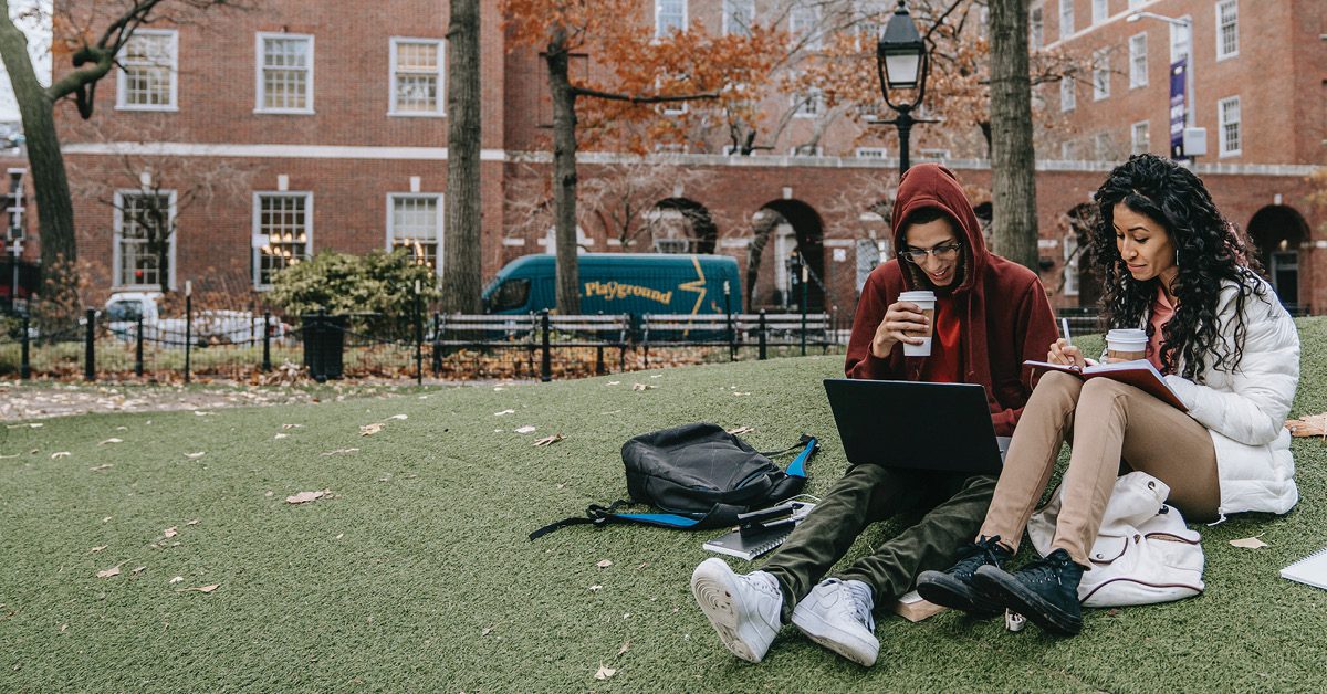 Two people sitting on the grass on a college campus looking at a laptop.