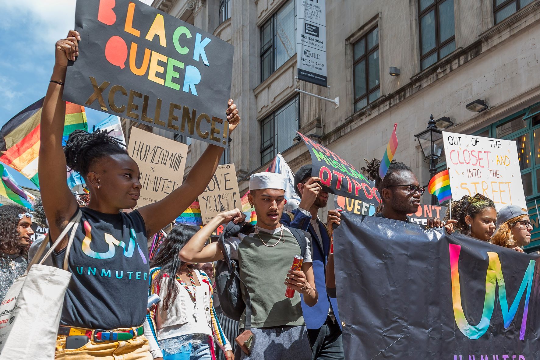 A group walking during a Pride event in Birmingham, UK. They are holding signs that read "Black Queer Excellence" and "Out of the Closet and into the Street."