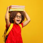 A young child wearing glasses and a red dress with a yellow backpack in front of a bright yellow background. They are holding books on top of their head and smiling enthusiastically with a big open mouthed smile.