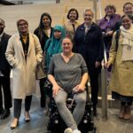 A picture of Seattle Disability rights commissions with delegates from Indonesia. They are standing in front of a wide wooden staircase. The person in front is in a motorized wheelchair.