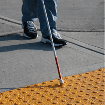 A person using a white cane approaches truncated domes as they cross the street.