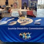 A member of the Seattle Disability Commission tabling at an event. They are holding up a shirt that defines the term disability.