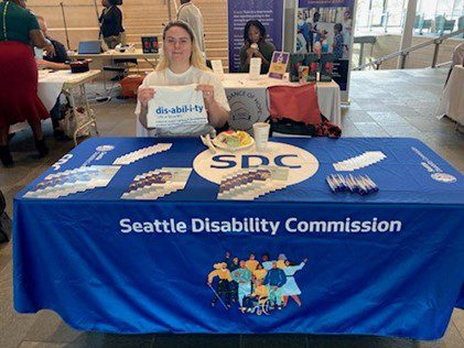 A member of the Seattle Disability Commission tabling at an event. They are holding up a shirt that defines the term disability.