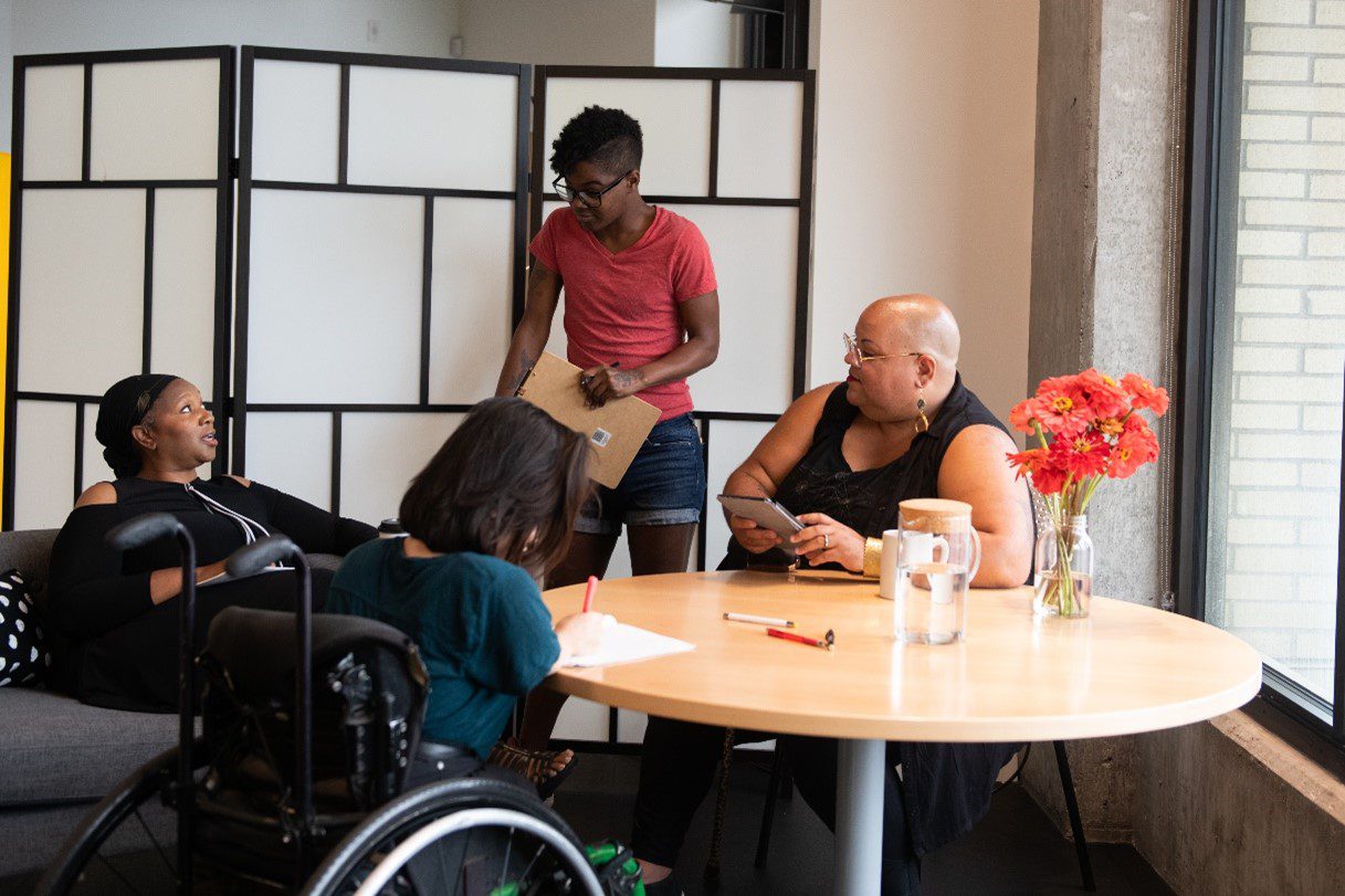Four people of color with disabilities gather around a table during a meeting.