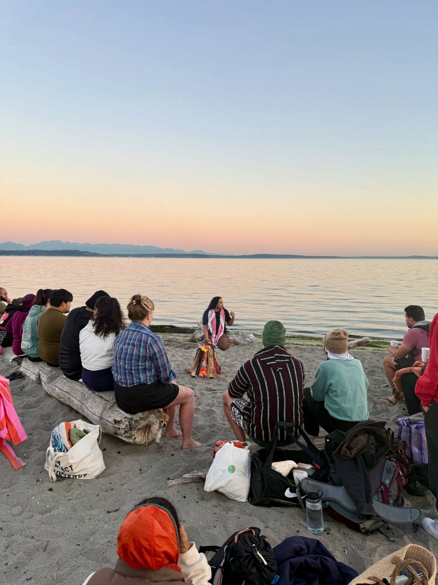 A large group of people gathered on the beach by a fire. 
